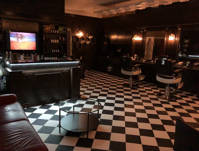 Dunkler Barberraum mit Lederlounge, warmem Licht und Checkerboard-Boden im Galaxy Barbershop Zürich